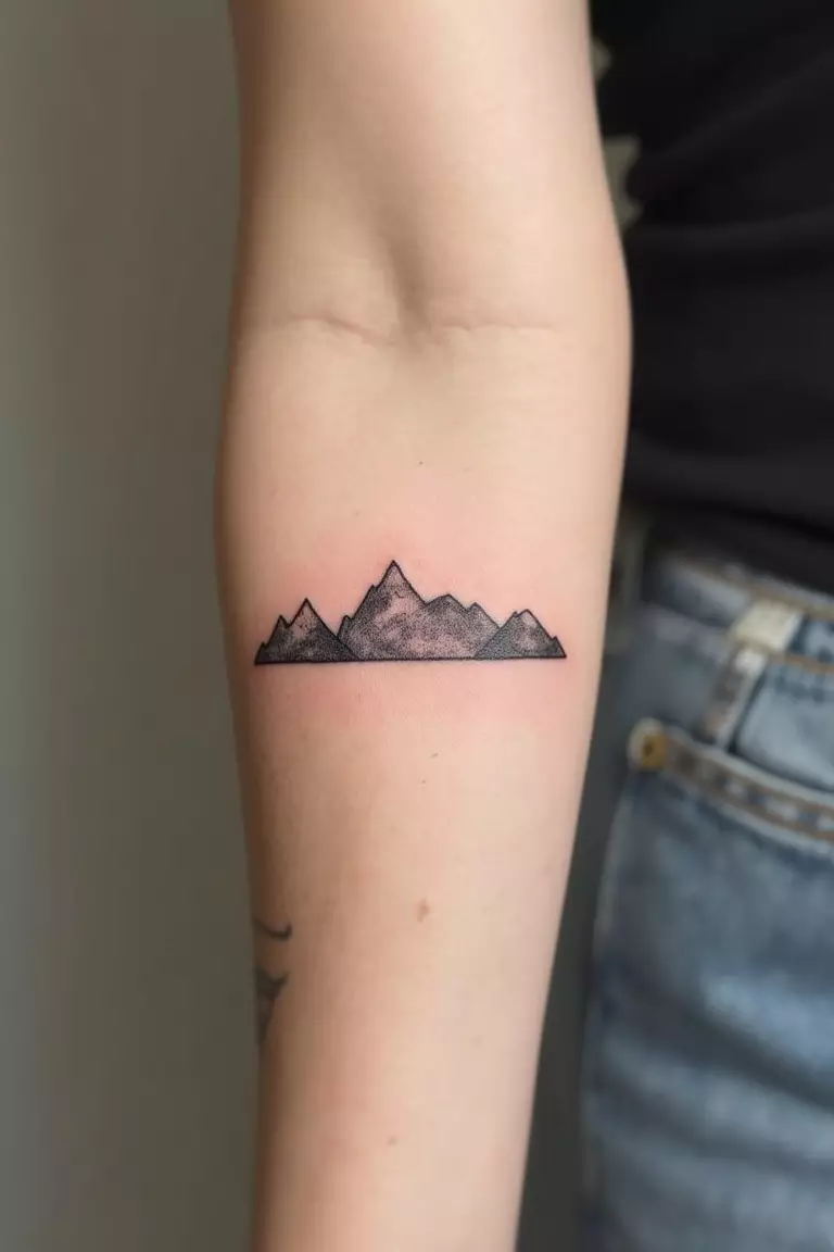 A close-up shot of a woman's arm with a black ink tattoo of a minimalist mountain range outline.
