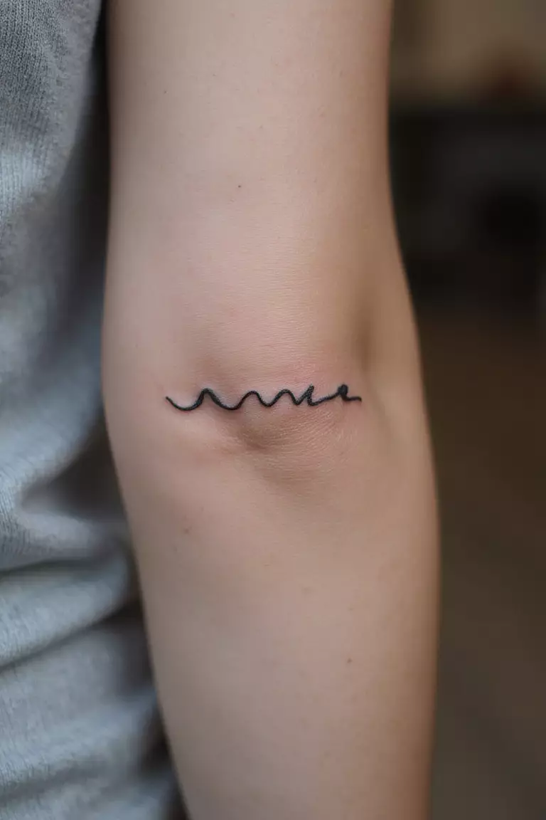 A close up shot of woman's arm with a black ink tattoo of a simple single line wave.