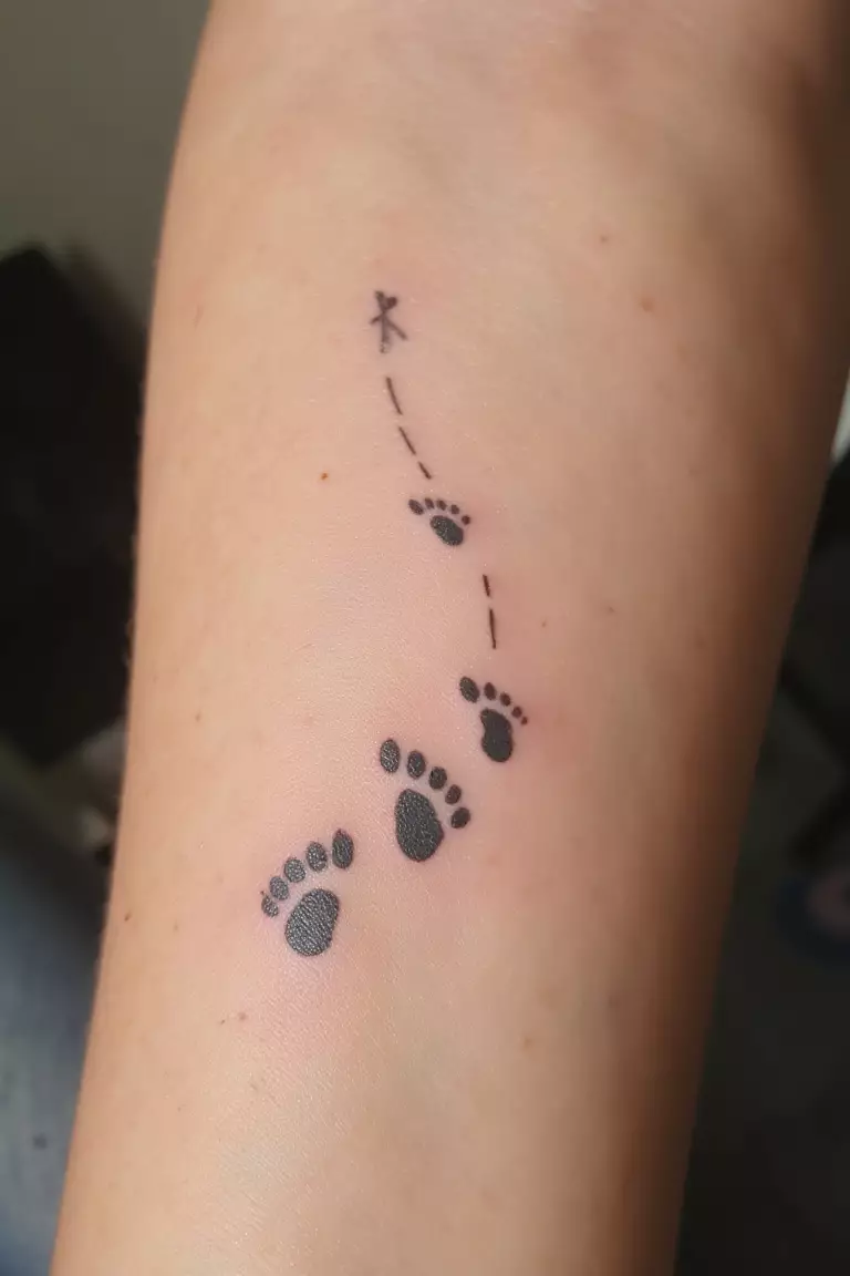 A close-up photo of a woman's arm showing her black ink tiny dog footprints trailing in a line tattoo