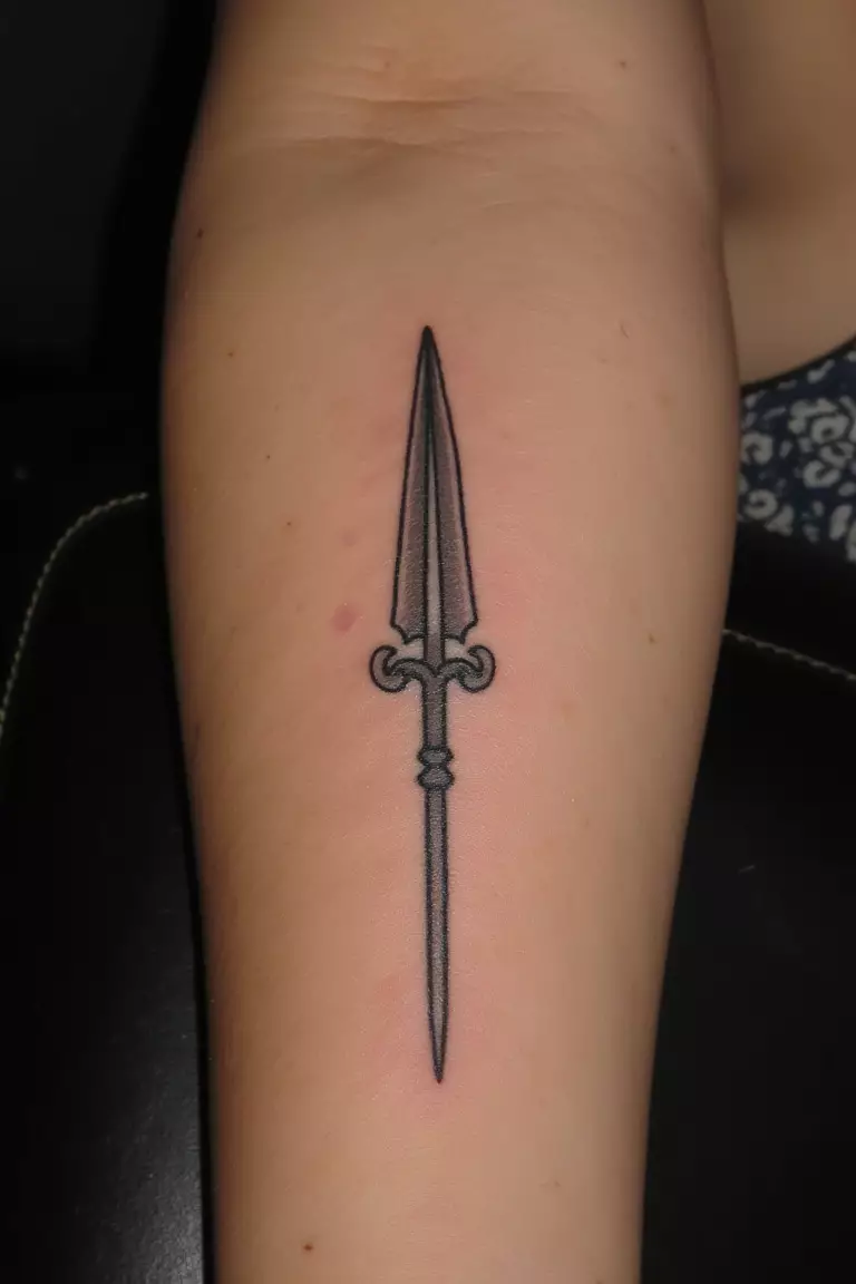 A close up shot of woman's arm with a black ink tattoo of a small spear representing Ares.