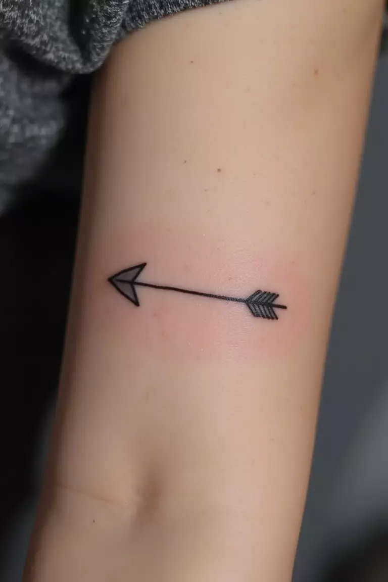 A close-up shot of a woman's arm with a black ink tattoo of a single arrow pointing forward.