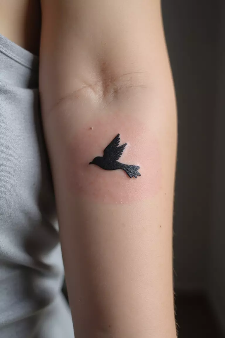 A close up shot of woman's arm with a black ink tattoo of a tiny flying bird silhouette.