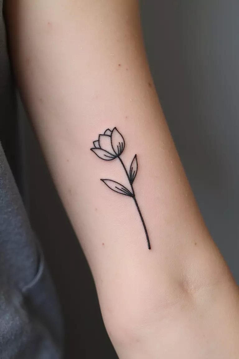 A close-up shot of a woman's arm with a black ink tattoo of a single stem minimalist line art flower, like a tulip or daisy.