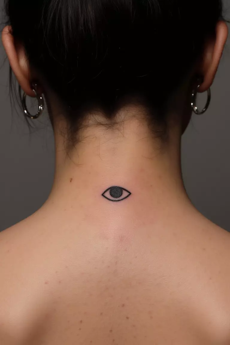 A close up shot of woman's neck from the back with a black ink tiny evil eye tattoo  