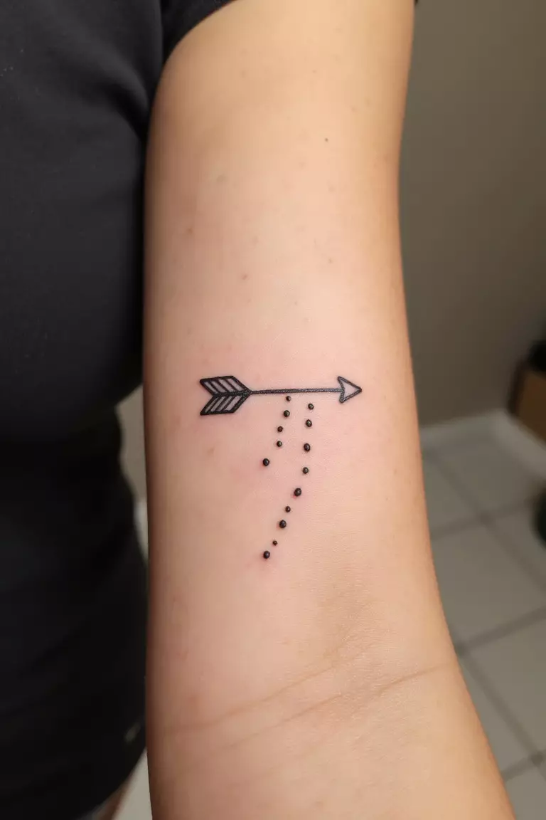 A close up shot of woman's arm with a black ink minimal arrow with dots tattoo, simple arrow accompanied by a trail of dots