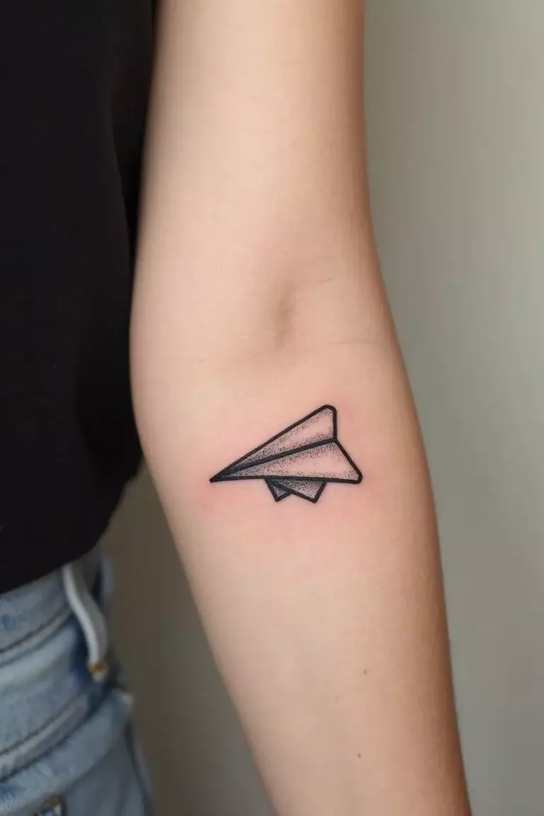 A close-up shot of a woman's arm with a black ink tattoo of a simple line art paper airplane.