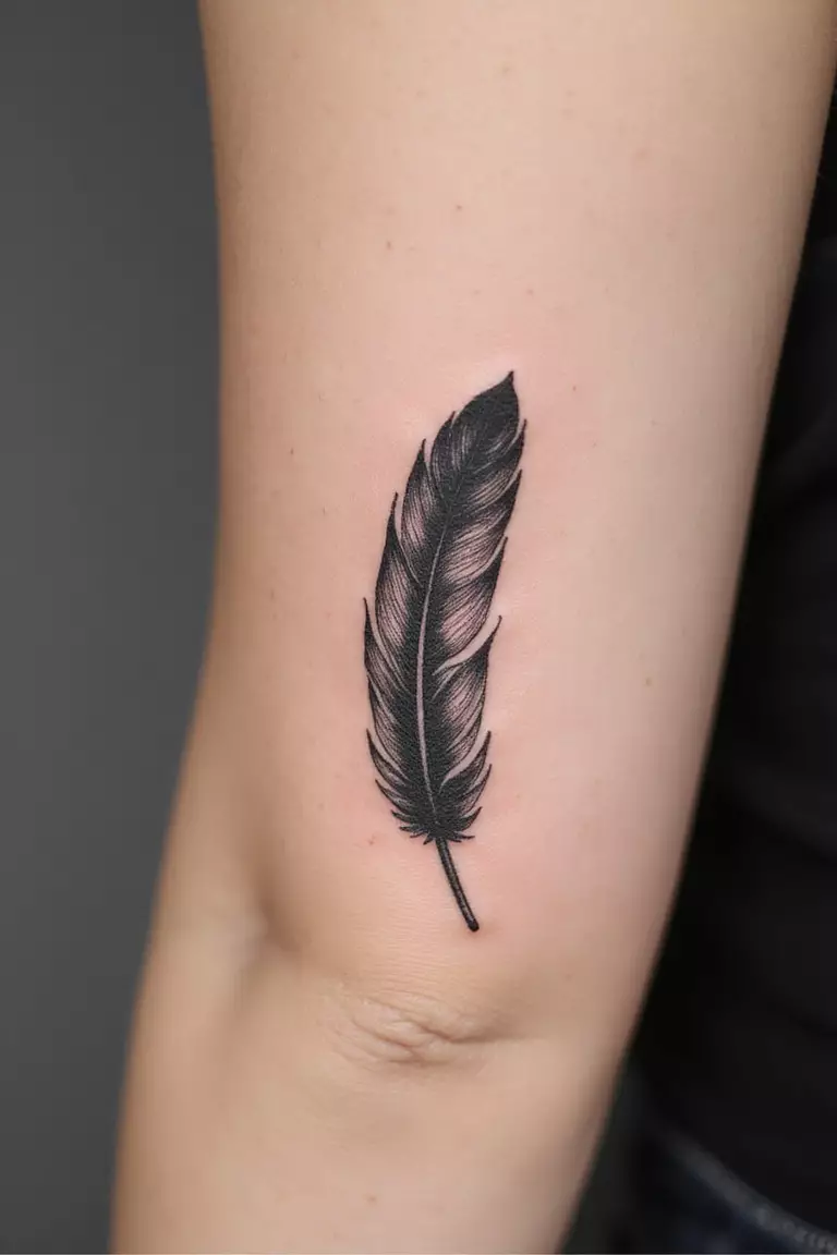 A close-up shot of a woman's arm with a black ink tattoo of a single, detailed feather seemingly floating down.
