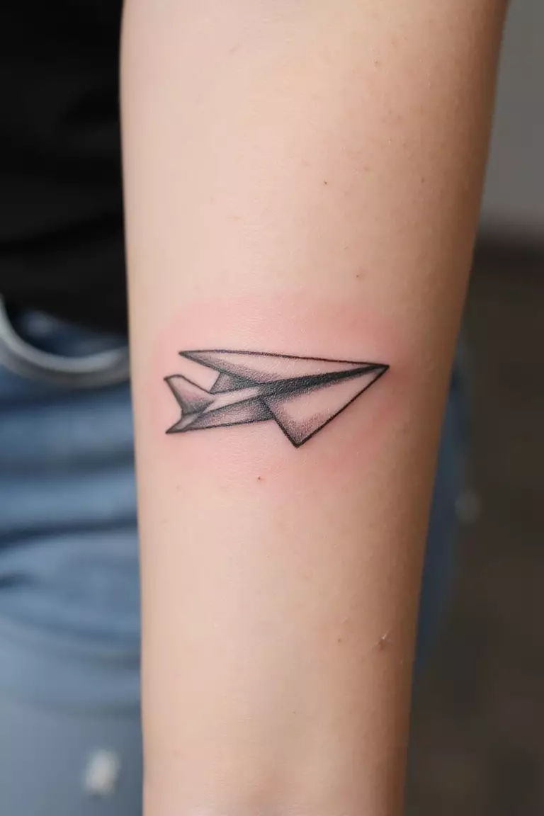A close up shot of woman's arm with a black ink tattoo of a simple paper airplane outline.