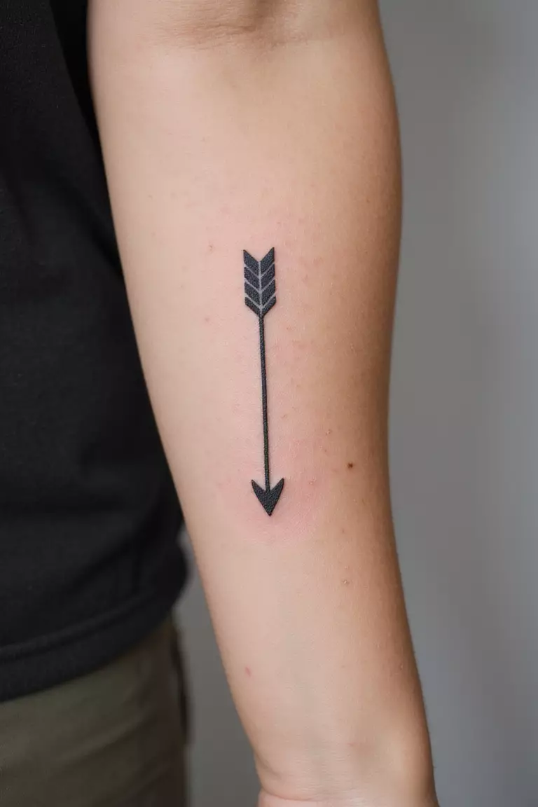 A close-up shot of a woman's arm with a black ink tattoo of a simple, thin black arrow.