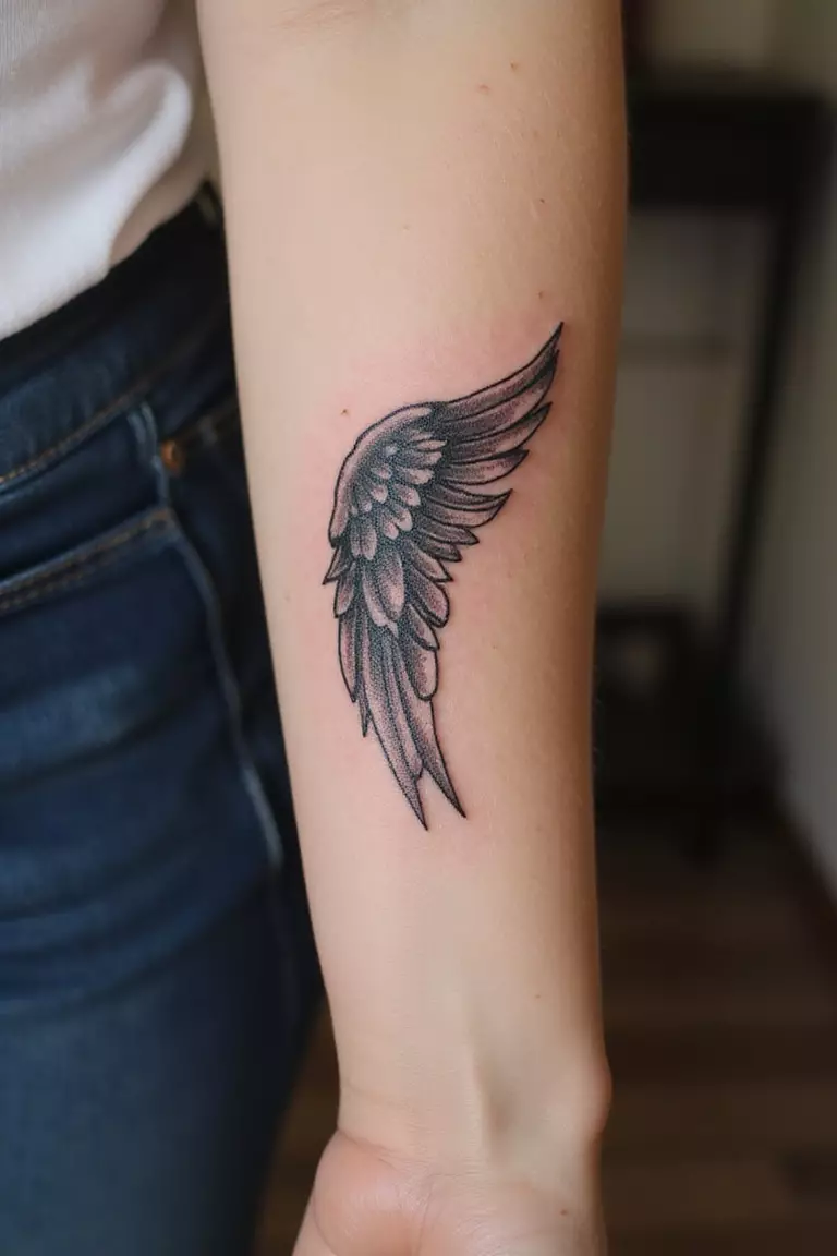 Small Angel Wing A close-up shot of a woman's arm with a black ink tattoo of a small angel wing.