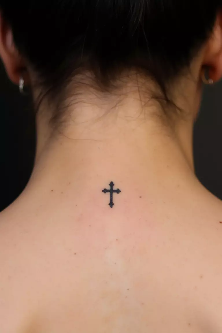 A close up shot of woman's neck from the back with a black ink tiny cross tattoo  