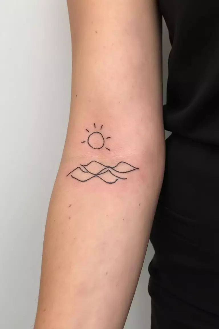 A close up shot of woman's arm with a black ink tattoo featuring minimal wave lines with a small outline of sun, perfect summer-themed design.