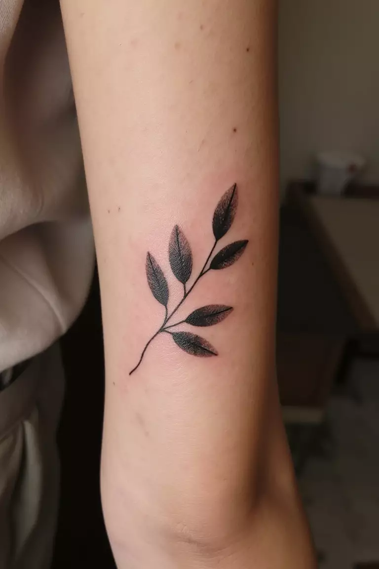A close up shot of woman's arm with a black ink olive branch featuring a subtle watercolor effect tattoo  