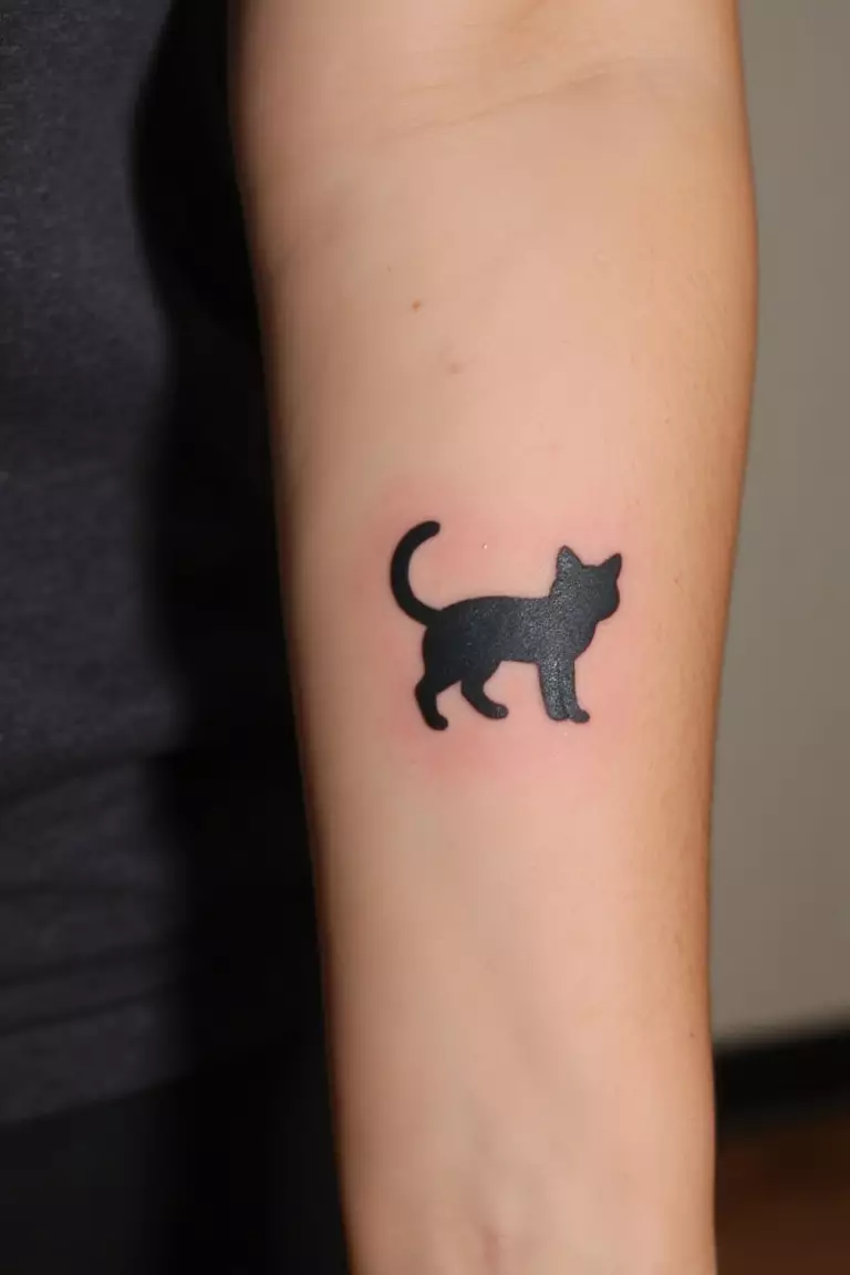 A close up shot of woman's arm with a black ink tattoo of a simple black cat silhouette.