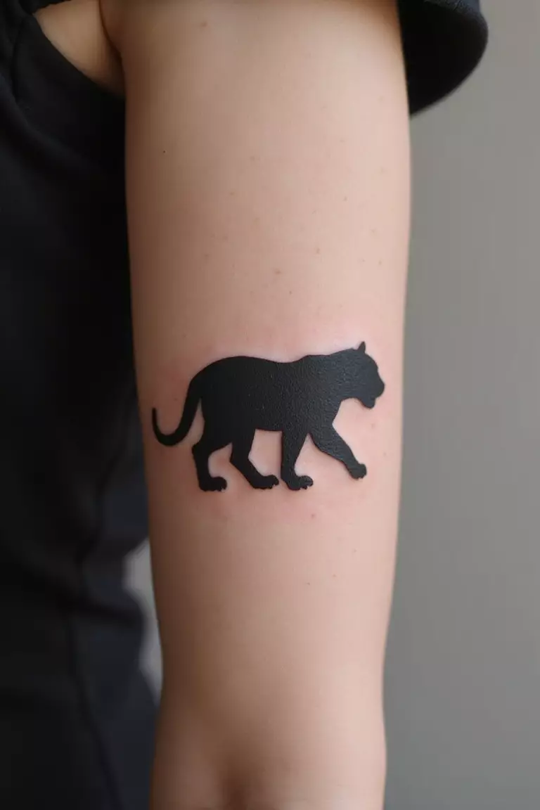 Tiger Silhouette Style A close-up shot of a woman's arm with a black ink tattoo of a solid black silhouette of a walking or crouching tiger.