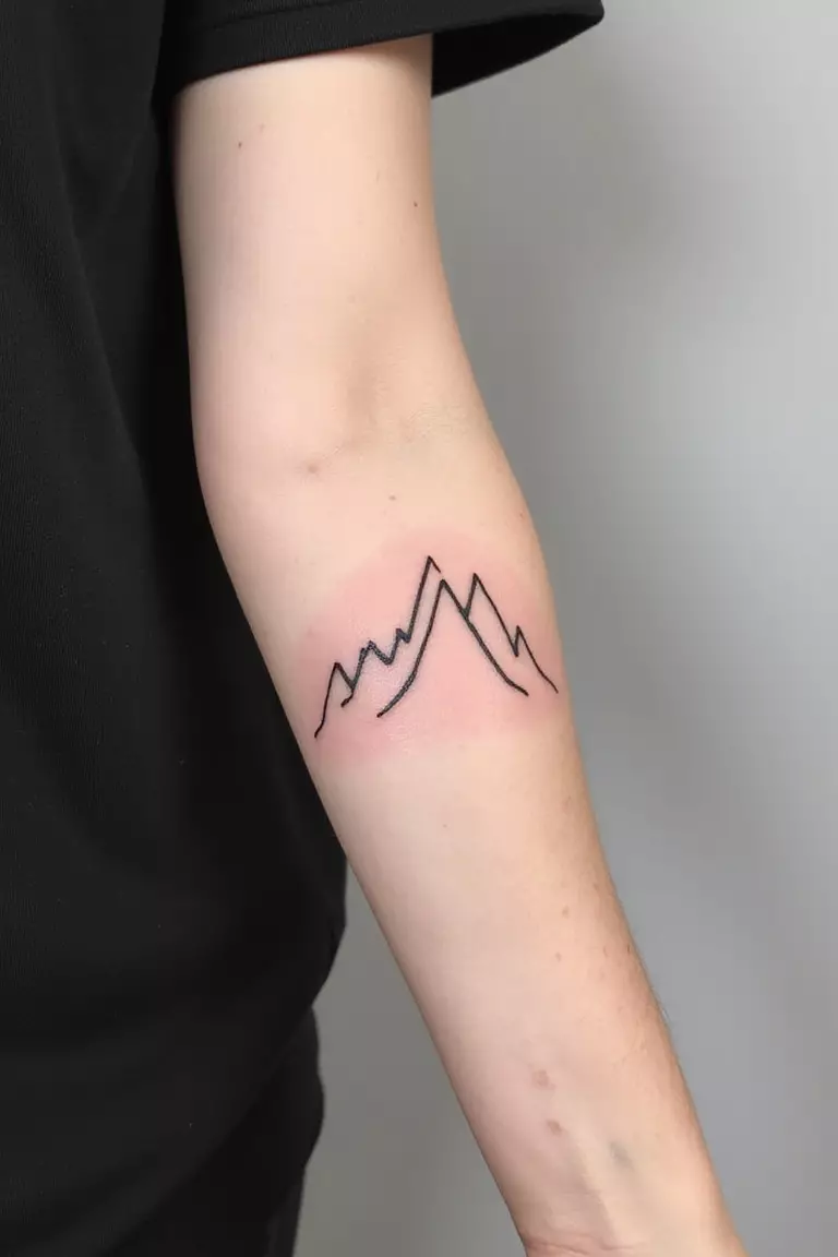 A close-up shot of a woman's arm with a black ink tattoo of a minimalist mountain range outline, simple peaks.