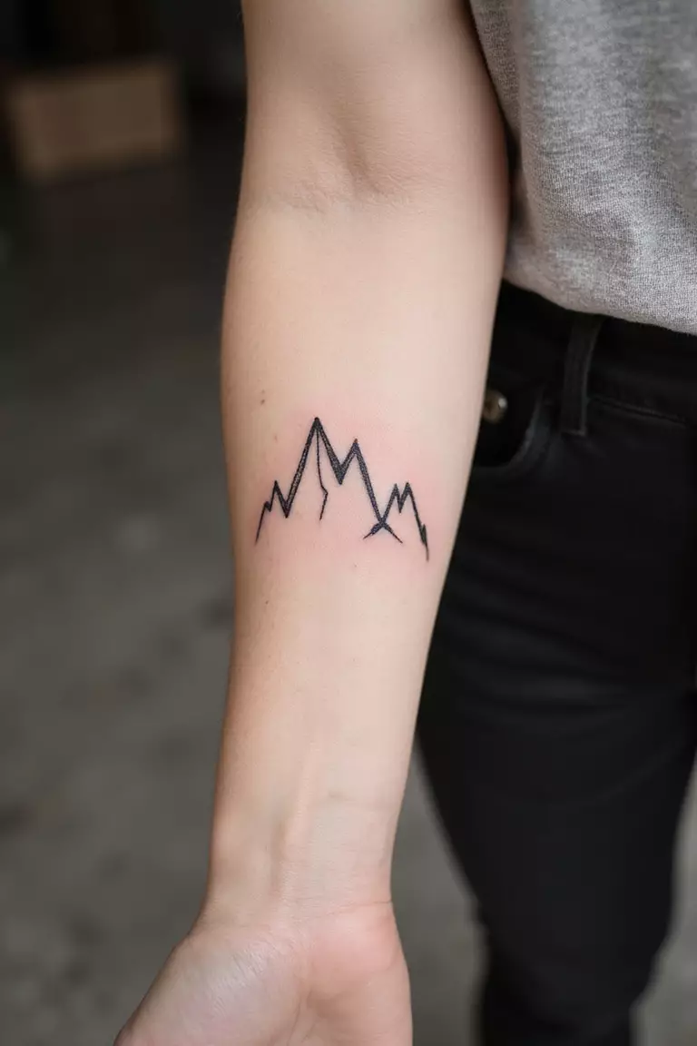 Mountain Range A close-up shot of a woman's arm with a black ink tattoo of a minimalist mountain range outline.