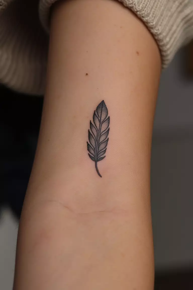 A close up shot of woman's arm with a black ink small feather wrist tattoo, dainty design perfectly positioned on inner wrist