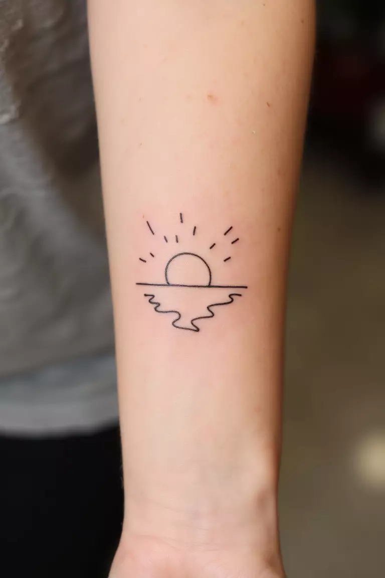 A close up shot of woman's arm with a black ink tattoo showing a minimal line outline of a sun setting over reflected water, tranquil beach-inspired design.