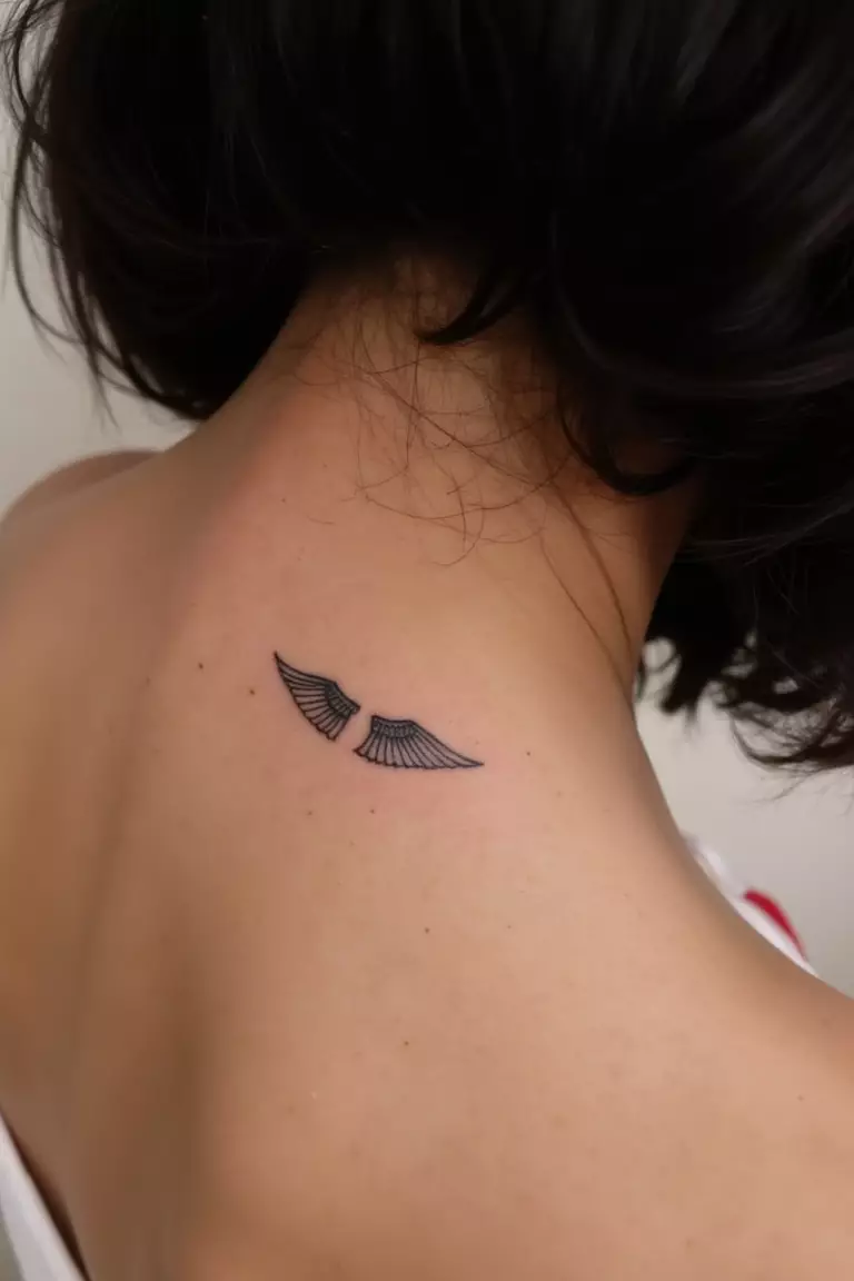 A close up shot of woman's neck from the back with a black ink tiny wings tattoo  